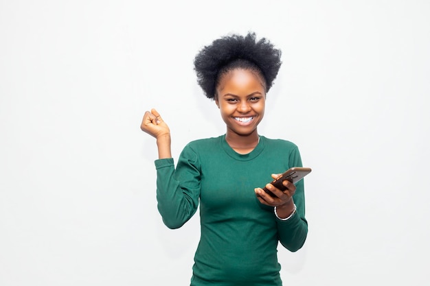 Confiante jovem africana segurando o telefone celular e olhando para ele com um sorriso em pé contra o fundo branco do estúdio