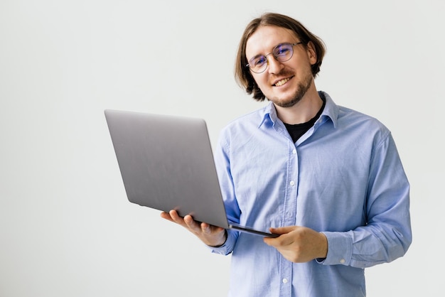 Confiante jovem bonito na camisa, segurando o laptop e sorrindo em pé contra um fundo branco