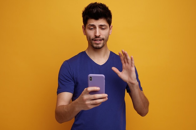 Confiante jovem bonito segurando e olhando para seu celular fazendo oi gesto em fundo amarelo