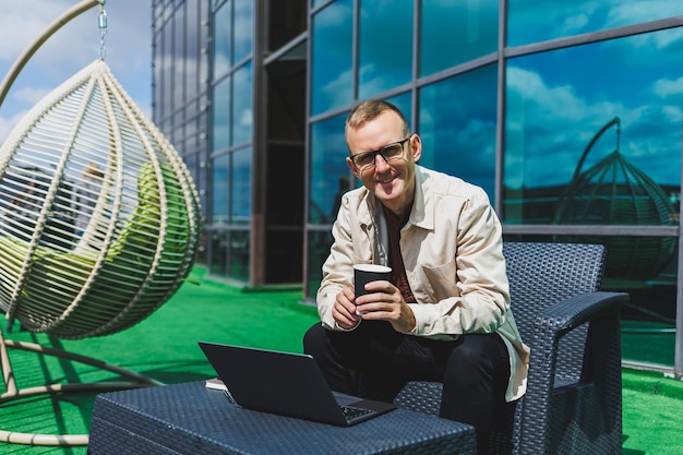 Confiante jovem empresário vestido com roupa formal e óculos elegantes chama um smartphone enquanto está sentado com café no terraço de um prédio de escritórios moderno com uma parede de vidro