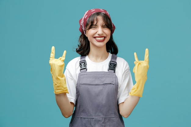 Confiante jovem limpador feminino usando bandana uniforme e luvas de borracha, olhando para a câmera mostrando sinal de rock com as duas mãos isoladas em fundo azul