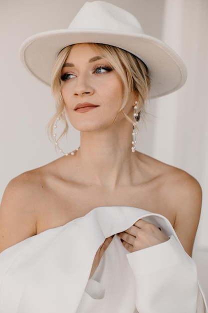 Confiante jovem loira sorrindo olhando para a câmera isolada no fundo branco Retrato de estúdio de uma mulher amigável bem-sucedida de terno branco e chapéu posando sobre parede branca