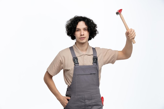 Confiante jovem trabalhador da construção civil vestindo uniforme olhando para a câmera levantando o martelo enquanto mantém a mão no bolso com chave de fenda no bolso isolado no fundo branco