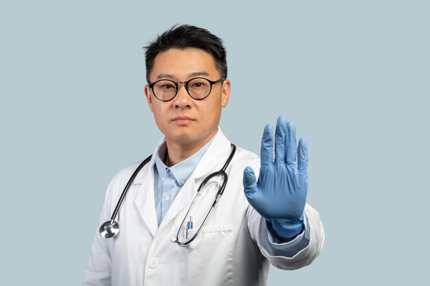 Confiante médico chinês de meia-idade sério em luvas e óculos de proteção de jaleco branco mostra sinal de pare