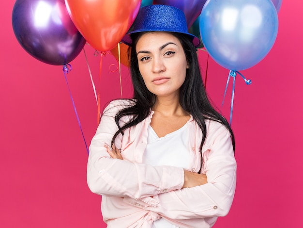 Confianza hermosa joven vistiendo gorro de fiesta de pie delante de globos cruzando las manos