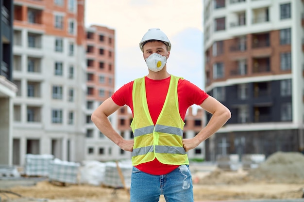 Confianza. Hombre confiado en casco de máscara protectora y chaleco brillante sosteniendo sus manos en el cinturón de pie en el sitio de construcción mirando a la cámara durante el día