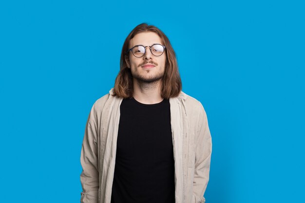 Confianza hombre de pelo largo con barba está mirando a la cámara con gafas y camisa en una pared azul del estudio