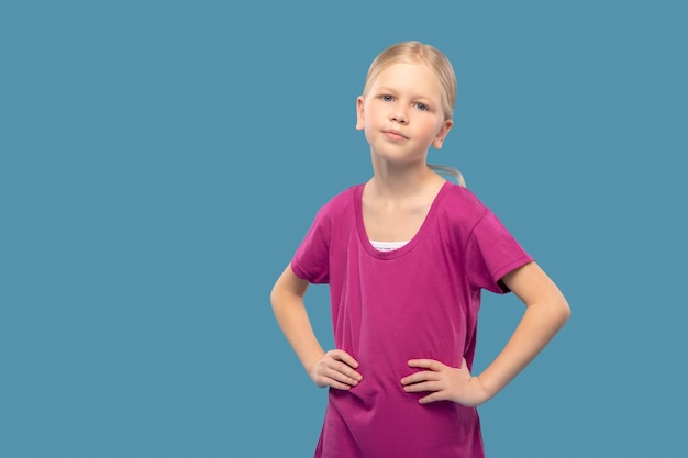 Confianza. Linda chica de escuela primaria rubia en camiseta deportiva brillante posando con las manos en el cinturón