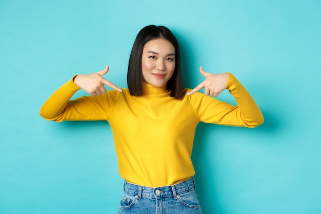 Confianza mujer asiática en elegante suéter apuntando con el dedo al logo en el centro, sonriendo asertiva a la cámara, de pie sobre fondo azul.