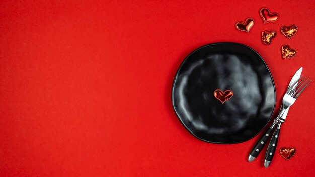 Configuração de dia dos namorados com garfo, faca, prato e corações em fundo vermelho. Vista superior, espaço de texto