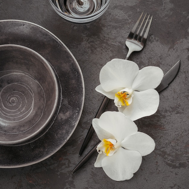 Configuração de mesa de elegância com guardanapo de malha cinza, talheres, pratos de cerâmica, copos e flores de orquídea brancas na mesa escura. Decoração de mesa moderna de férias. Jantar romântico.