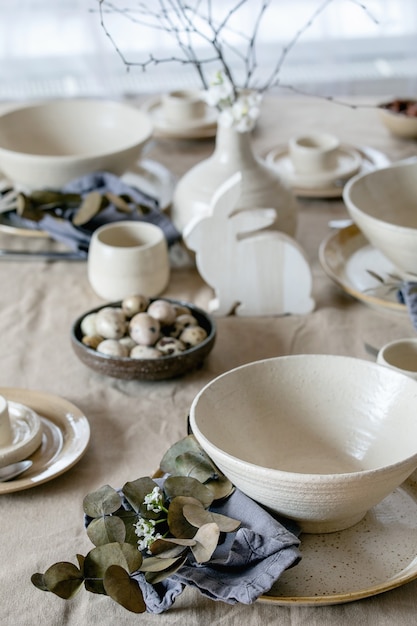 Configuração de mesa de Páscoa rústica com talheres de cerâmica artesanais vazios, pratos e tigelas, decorações de coelho de Páscoa, ramos em um vaso na toalha de linho na frente da janela. Jantar de Páscoa.