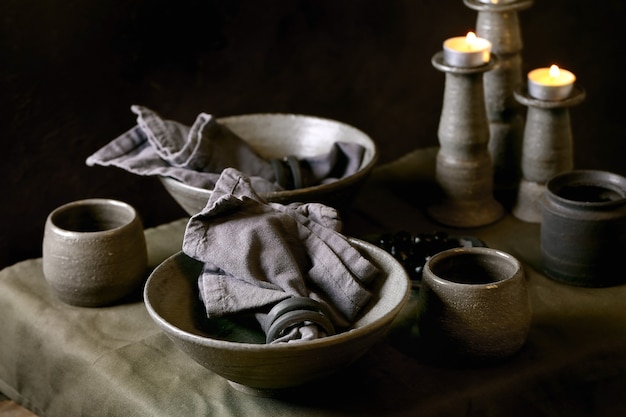 Configuração de mesa rústica com talheres de cerâmica artesanais vazios, tigelas e xícaras cinzas ásperas, velas acesas na toalha de linho. fundo escuro