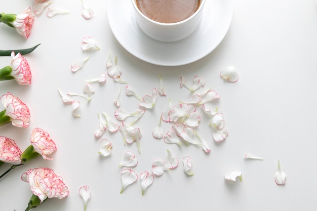Configuração lisa do café no copo branco com as flores e as pétalas cor-de-rosa do cravo na mesa branca.