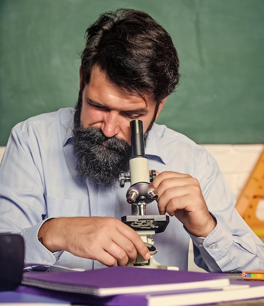 Configurar microscopio Profesor sentarse escritorio con microscopio Hombre barbudo hipster aula pizarra fondo ocupado con observación biológica Profesor de escuela mirando microscopio Investigación fascinante