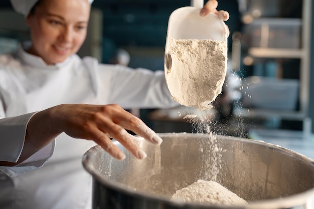 Confitería profesional con uniforme blanco poniendo harina en un gran tazón de metal de pie cerca de la mesa en la confitería. Vista de primer plano, enfoque selectivo