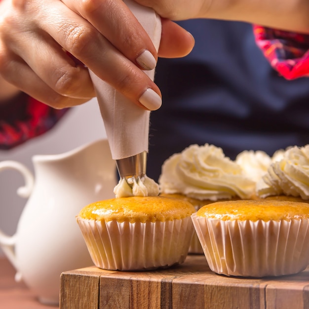 Confitero femenino en un delantal azul y una camisa roja a cuadros aplica crema a los cupcakes