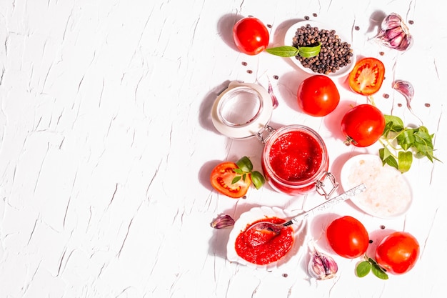 Confitura de tomate, mermelada, chutney, salsa en frasco de vidrio. Concepto de conservación casera. Tomates frescos, especias, ajo, verduras, sal marina. Luz dura moderna, sombra oscura, fondo de masilla blanca, vista superior