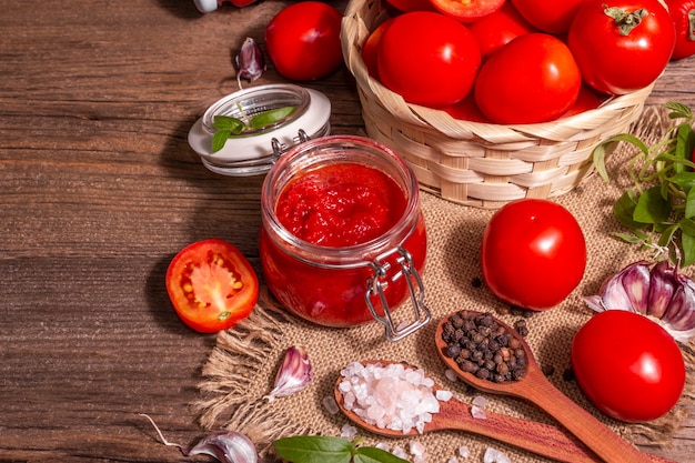 Confiture de tomate, geléia, chutney, molho em uma jarra de vidro. Conceito de preservação caseiro. Tomates frescos, especiarias, alho, verduras, sal marinho. Luz dura moderna, sombra escura, mesa de madeira vintage, espaço de cópia