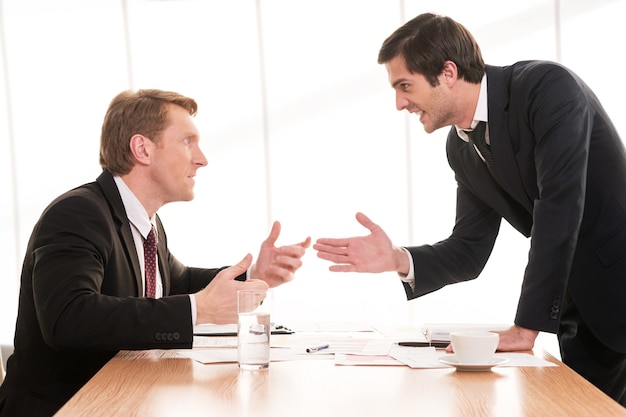 Conflicto comercial. Dos hombres jóvenes en ropa formal discutiendo y gesticulando mientras están sentados en la mesa