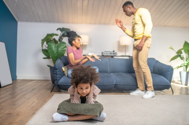 Conflicto. Padres jóvenes de piel oscura jurando nerviosamente en la parte trasera de la habitación en el sofá y su pequeña hija infeliz con los oídos cerrados sentados en el suelo