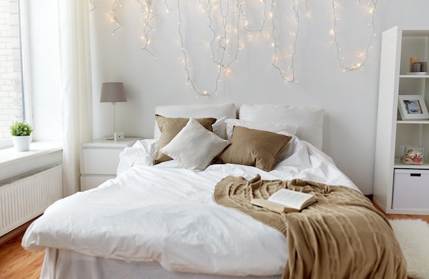 conforto, conforto, interior e conceito de férias - quarto aconchegante com cama e luzes de guirlandas de Natal em casa