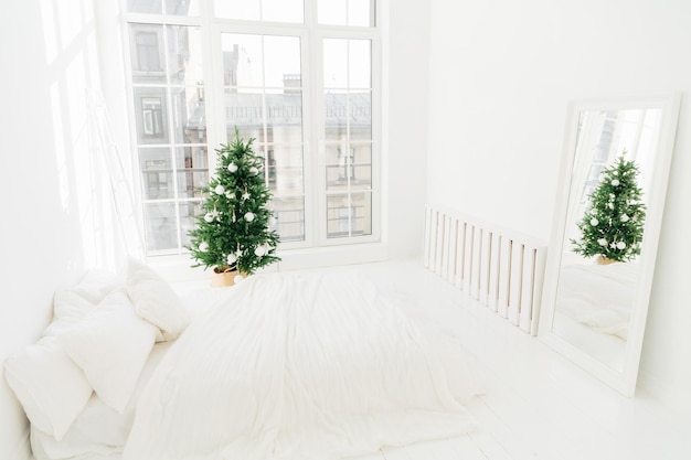 Conforto em casa e conceito de véspera de Natal Quarto com espelho de cama macia branca grande janela para luz entrando no quarto decorado Árvore de Ano Novo e escada Decoração de férias