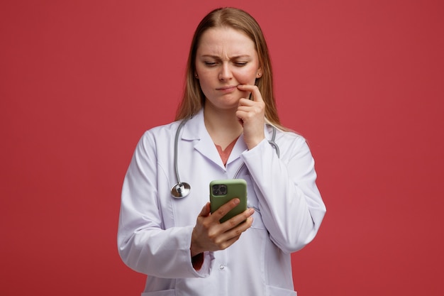 Confundida joven doctora rubia vistiendo bata médica y estetoscopio alrededor del cuello sosteniendo y mirando el teléfono móvil manteniendo el dedo en la esquina de la boca