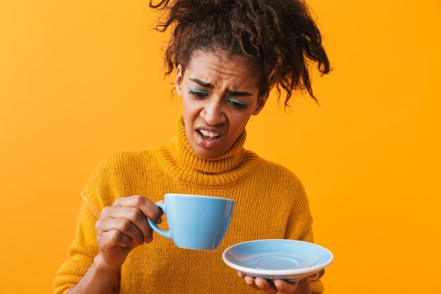 Confundida mujer africana vistiendo suéter sosteniendo la taza en el platillo aislado