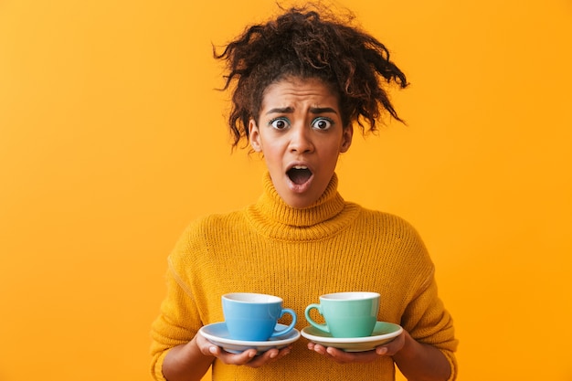 Confundida mujer africana vistiendo suéter sosteniendo tazas en platillos aislado