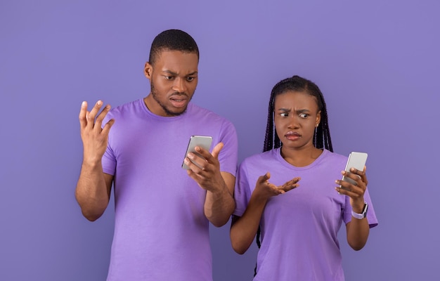 Confundida pareja afroamericana milenaria sosteniendo usando sus teléfonos celulares