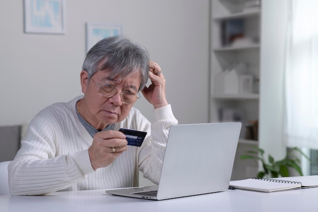 Confundido, un anciano asiático jubilado, estresado, preocupado, triste, frustrado, hombre, que tiene problemas para pagar, comprar pagos en línea con crédito, tarjeta bancaria bloqueada, computadora portátil, fraude en Internet