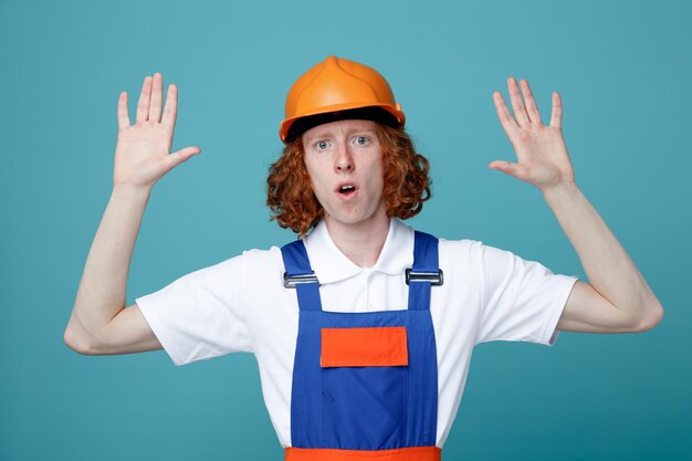 Confundido extendiendo las manos joven constructor hombre en uniforme aislado sobre fondo azul.