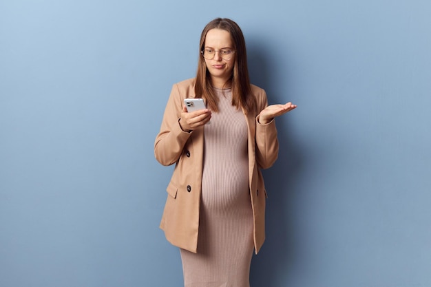 Confusa, desconcertada, joven adulta embarazada con vestido y chaqueta sosteniendo un teléfono móvil mirando la pantalla de su teléfono inteligente con una postura incierta aislada sobre un fondo azul