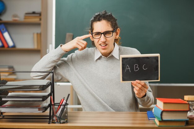 confuso colocando o dedo na cabeça jovem professor de óculos segurando o mini quadro-negro sentado na mesa com as ferramentas da escola na sala de aula