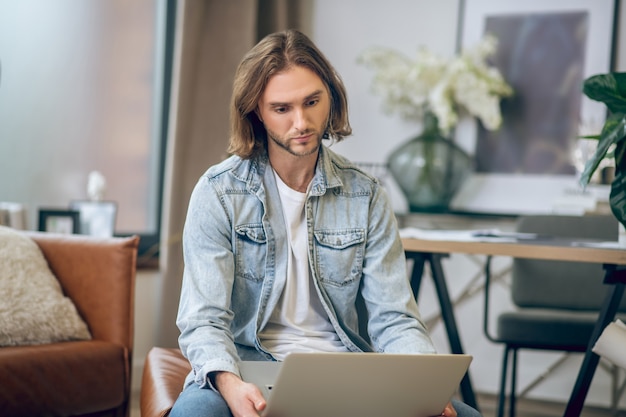 Confuso. Homem de camisa jeans trabalhando no laptop e parecendo confuso