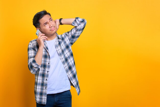 Confuso jovem asiático em camisa xadrez falando no celular olhando de lado no espaço de cópia isolado em fundo amarelo