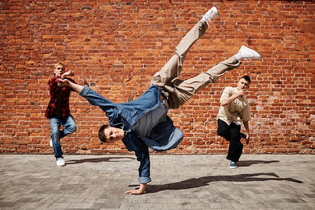 Congele o quadro do artista de breakdance masculino fazendo pose de parada de mão com a equipe contra a parede de tijolos ao ar livre