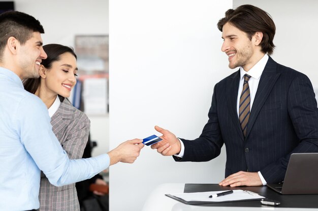 Cônjuges felizes comprando carro dando cartão de crédito de vendedor permanente no escritório de concessionária de automóveis. espaço vazio. vista lateral