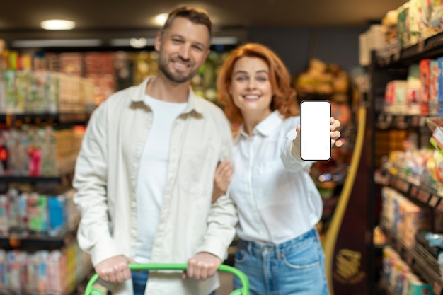 Cônjuges felizes de pé no supermercado e mostrando smartphones em branco comprando e recomendando aplicativos