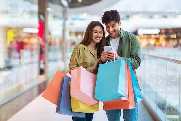 Cônjuges felizes fazendo compras e usando o celular em pé no shopping