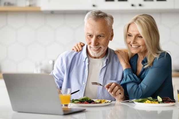 Cônjuges idosos sorridentes relaxando com laptop enquanto almoçam na cozinha