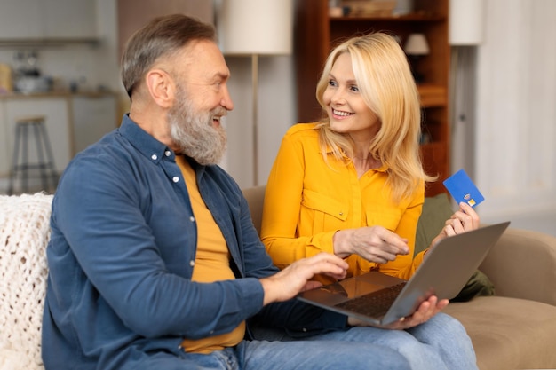 Cônjuges idosos usando cartão e laptop desfrutando de compras on-line em ambientes fechados