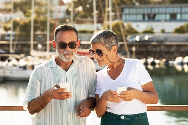 Cônjuges maduros usando telefones posando no cais da marina com veleiros