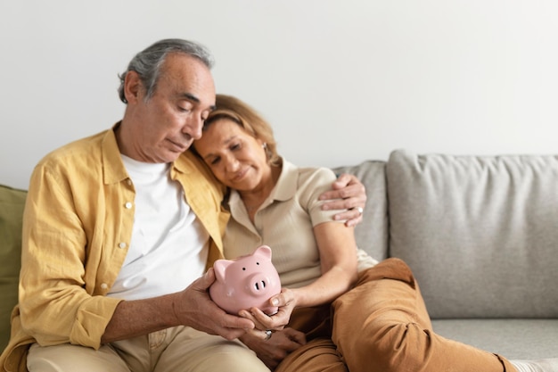 Cônjuges sênior de poupança de aposentadoria segurando cofrinho enquanto sentados juntos no sofá e se abraçando
