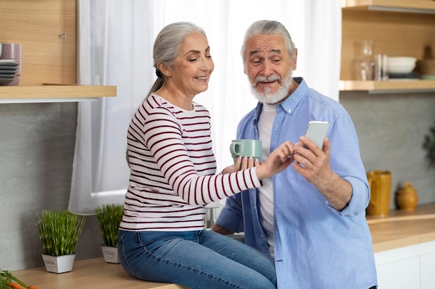 Cônjuges sênior felizes usando smartphone juntos enquanto relaxam na cozinha