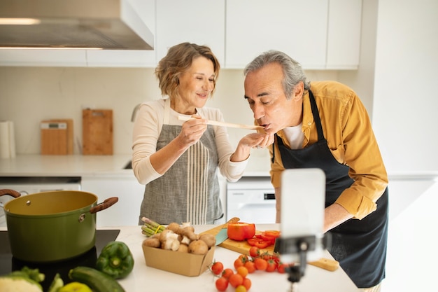 Cônjuges superiores positivos cozinhando jantar saudável e mulher alimentando o marido transmitindo e usando