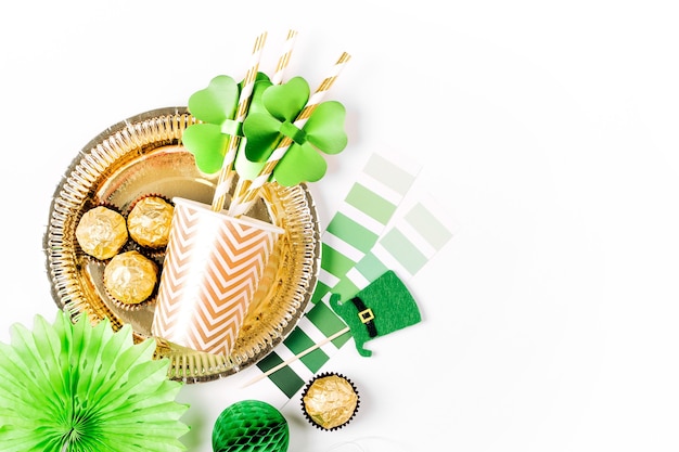 Conjunto de accesorios para la fiesta del Día de San Patricio. Decoraciones de papel verde y dorado, platos y vasos desechables, sombreros, hoja de trébol, confeti y dulces sobre fondo blanco. Vista plana endecha, superior.