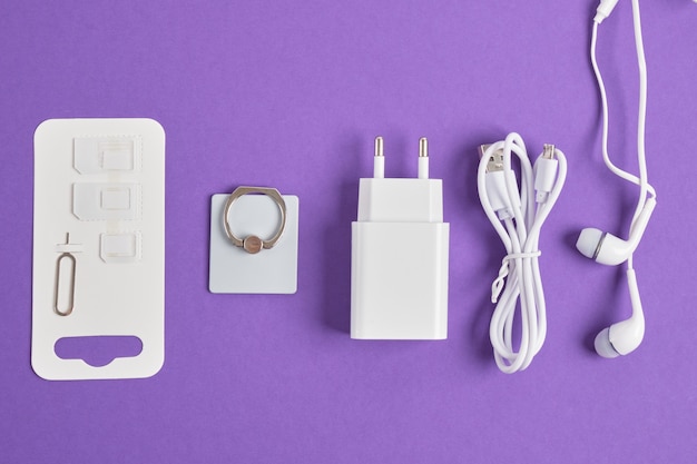 Un conjunto de accesorios para un teléfono inteligente, cargador, auriculares, un anillo, adaptadores para tarjetas SIM en una vista superior del espacio de copia de fondo púrpura