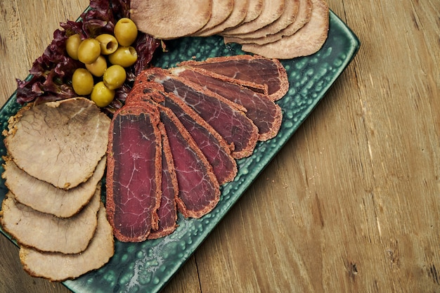 Conjunto de carne antipasti - lengua al horno en rodajas, jamón, cerdo hervido y basturma en un plato azul sobre una superficie de madera. Merienda cerveza